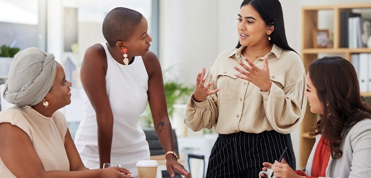 Four professional women engaged in a dynamic discussion around a table in a bright office.