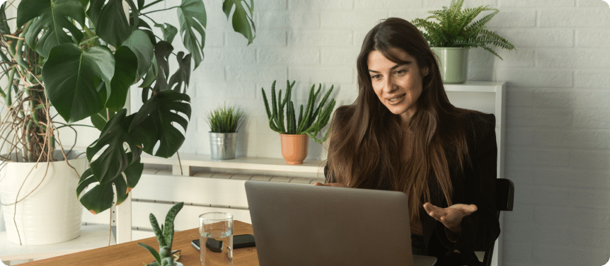 Woman on a video call from a plant-filled home office, gesturing as she speaks.