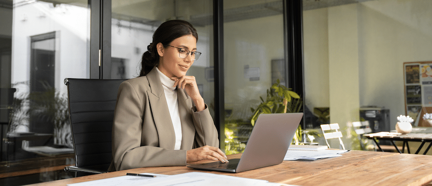 Professional woman in a blazer working on a laptop in a bright, modern office.