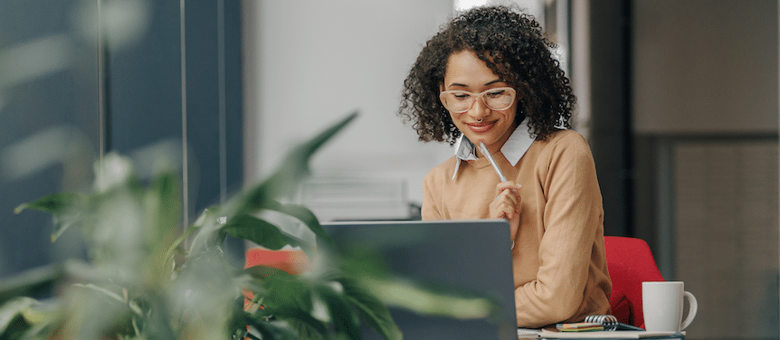 Professional woman with curly hair and glasses smiling at her laptop in a bright, plant-filled office.